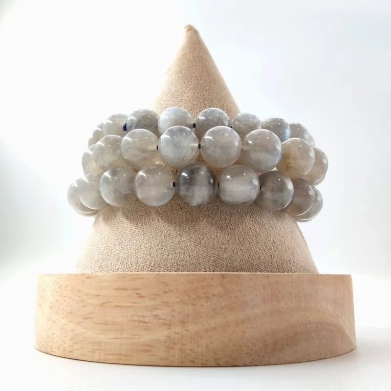 ette image présente trois bracelets de perles grises irisées, disposés sur un présentoir conique en tissu beige avec un socle en bois clair. Les perles sont des Labradorites, reconnaissables à leur teinte gris perle et à leurs reflets bleutés métalliques qui apparaissent selon l'angle de la lumière. Contrairement aux modèles précédents, ces pierres ont un aspect plus froid et minéral, parsemé de petites zones sombres et de structures internes feuilletées qui capturent l'éclat. L'ensemble est photographié sur un fond blanc neutre, soulignant la brillance satinée de chaque bille.