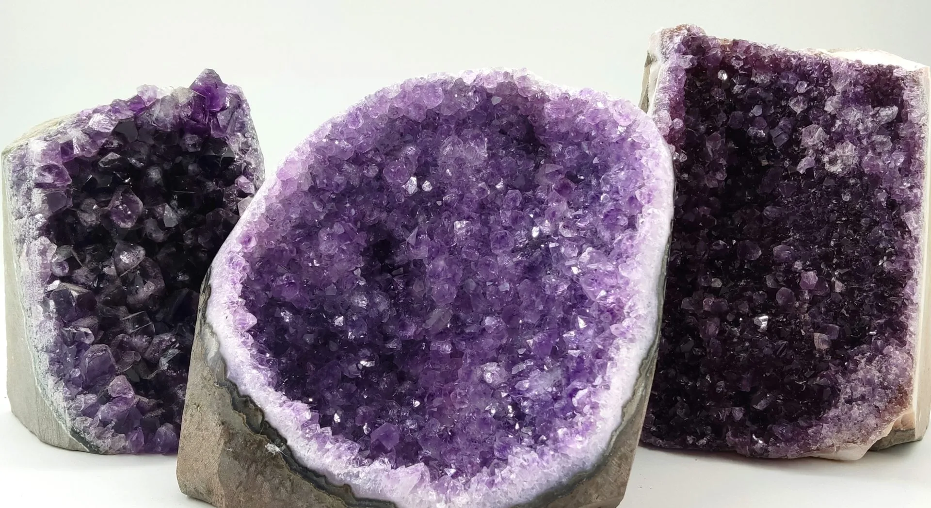 Trois géodes d’améthyste posées côte à côte, révélant leurs cristaux violets étincelants. La pierre du centre présente des nuances claires et lumineuses, tandis que celles à gauche et à droite affichent des tons plus profonds et intenses.