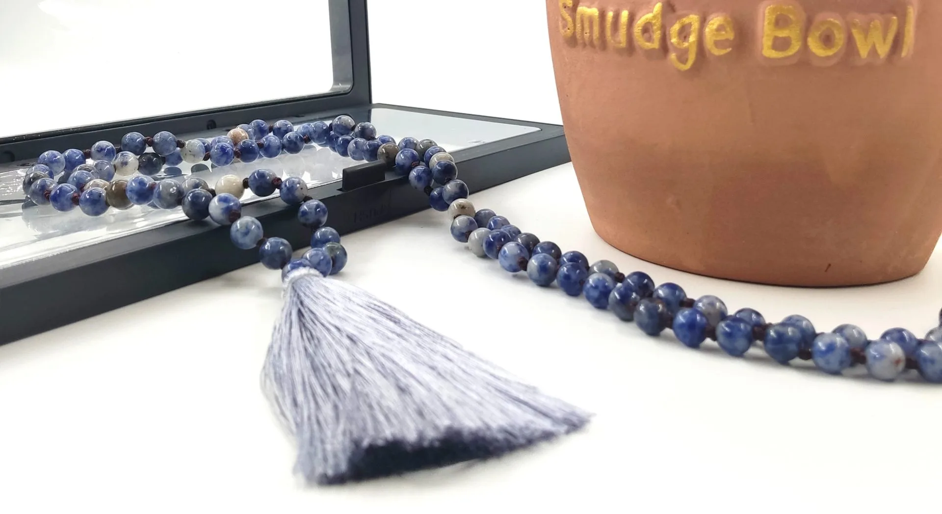 Perles rondes en pierre bleue, terminé par un pompon bleu clair, posé devant un miroir et à côté d’un bol marqué 'Smudge Bowl'.