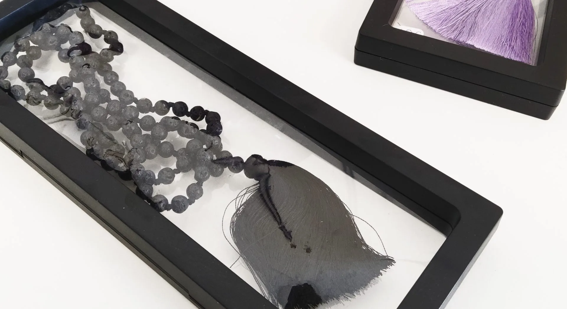 perles grises avec un pompon noir, présenté dans un cadre noir transparent, à côté d’un autre cadre contenant un pompon violet.
