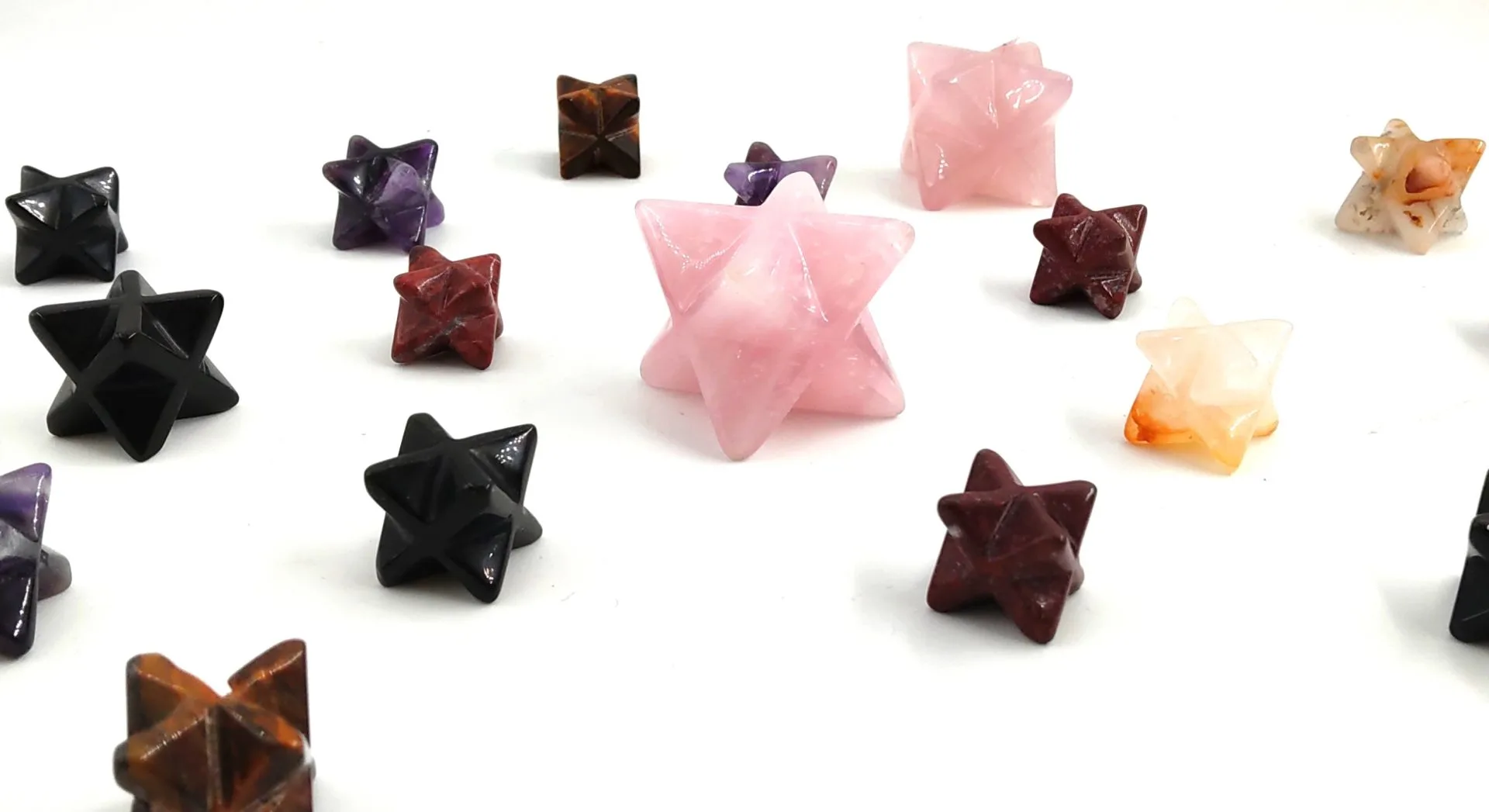 Collection de Merkabas géométrie sacrée en pierres naturelles polies, dont quartz rose, œil de tigre, jaspe rouge, améthyste et obsidienne noire, disposés sur fond blanc.