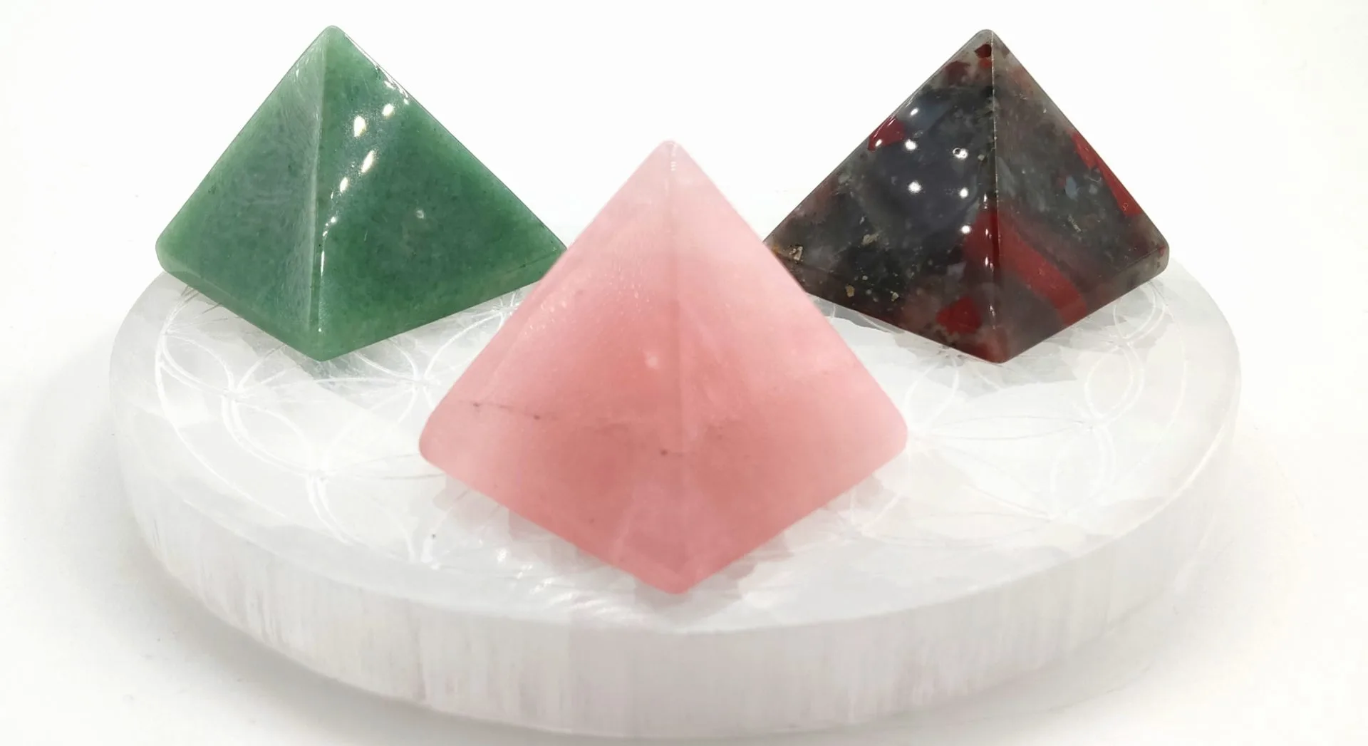 Trois pyramides géométrie sacrée en pierres naturelles — aventurine verte, quartz rose et jaspe rouge — posées sur une plaque ronde en sélénite gravée de la Fleur de Vie.