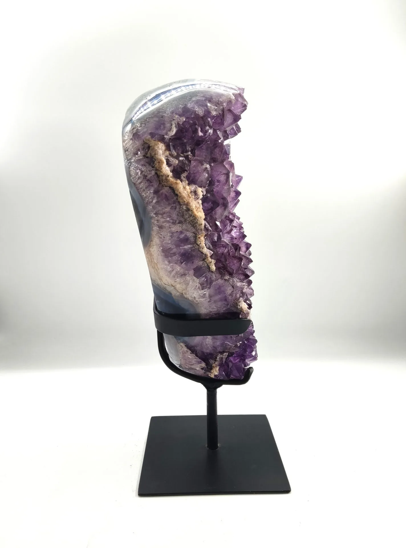 Coupe verticale de géode d’améthyste montée sur un support métallique noir. La tranche laisse apparaître une bordure cristallisée violette intense avec des couches minérales beiges et grises.