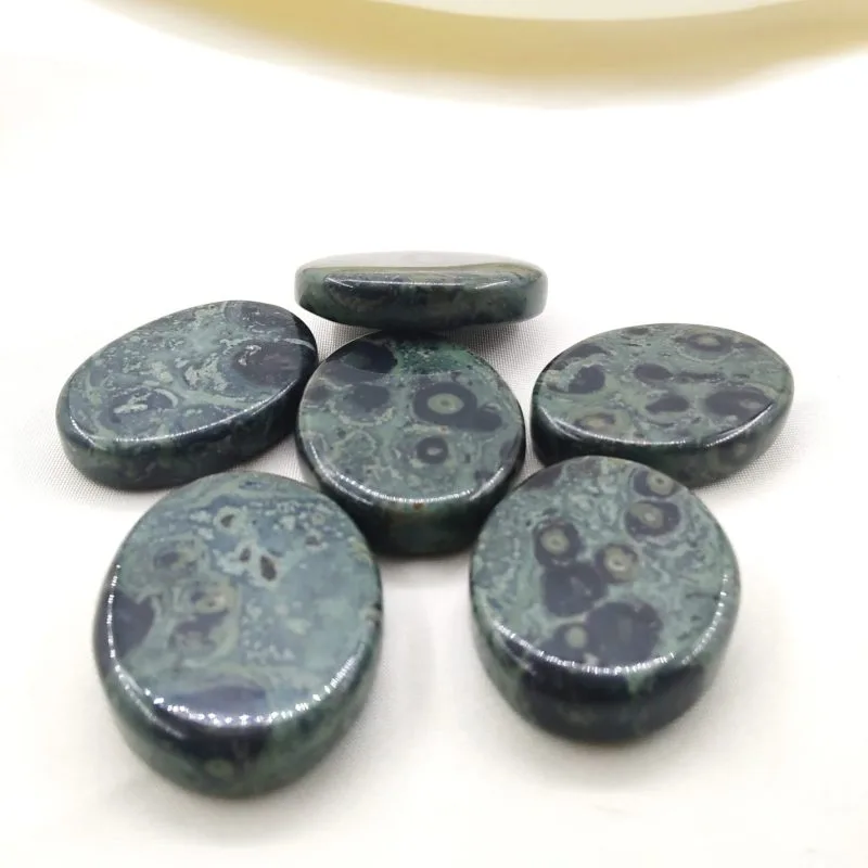 Six pierres polies en jaspe kambaba, aux motifs verts et noirs rappelant des cercles et volutes, posées sur un tissu blanc crème.