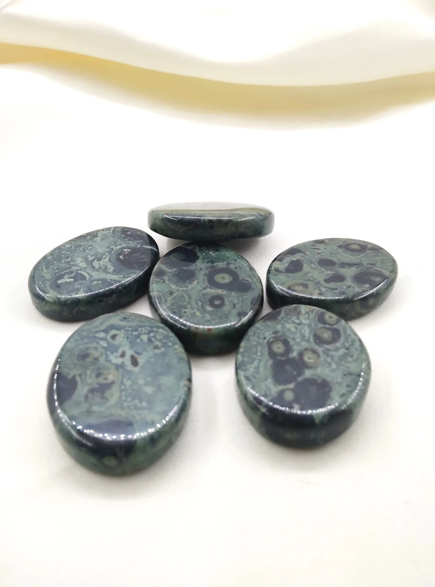 Six pierres polies en jaspe kambaba, aux motifs verts et noirs rappelant des cercles et volutes, posées sur un tissu blanc crème.