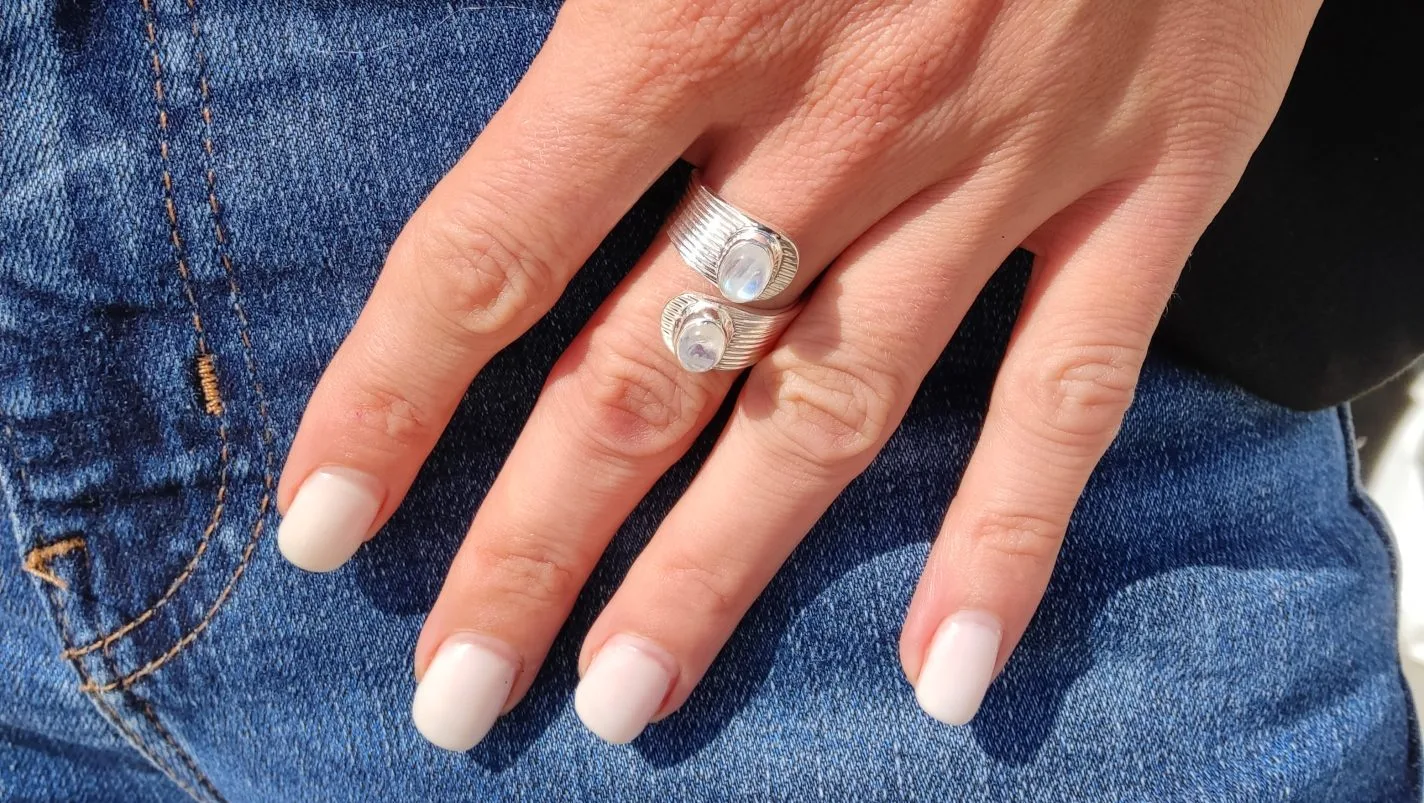 Main portant une bague en argent ajustable ornée de deux péristérite ovales blanches aux reflets irisés, photographiée en extérieur avec un jean et une veste noire.