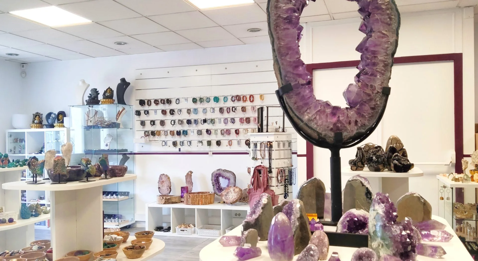 Intérieur d’une boutique de minéraux et lithothérapie, avec de grandes géodes d’améthyste, des pierres polies, des bijoux en pierres naturelles exposés sur des présentoirs blancs et un mur garni de bracelets colorés.