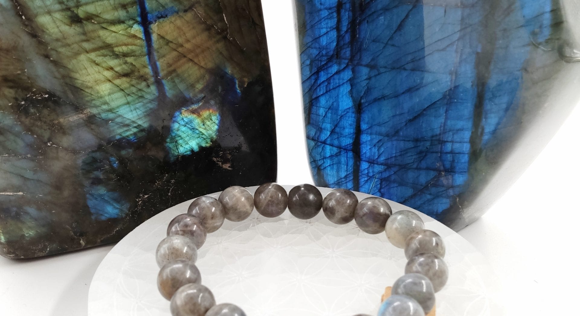 Bracelet en perles de labradorite posé sur un support décoré du motif Fleur de Vie, avec en arrière-plan deux grandes labradorites polies aux reflets bleu, vert et doré irisés.