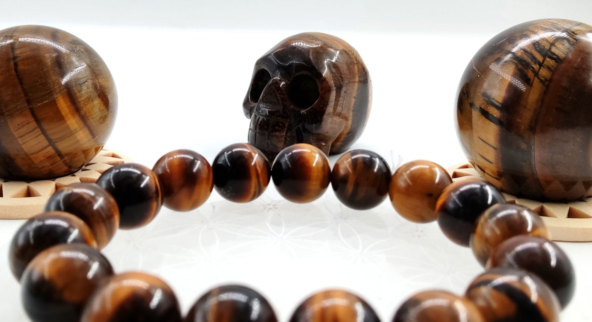 Bracelet en perles d’œil de tigre posé au premier plan, accompagné d’un crâne sculpté et de deux sphères polies en œil de tigre aux reflets chatoyants brun, or et caramel.