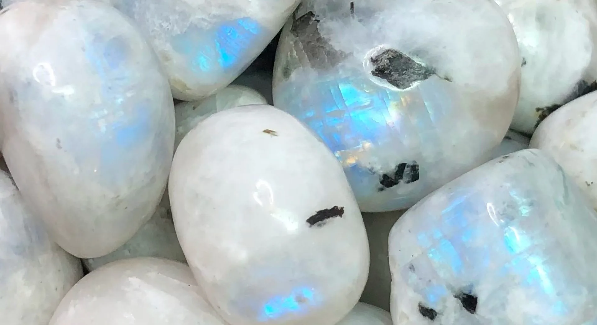 Pierres de lune polies, blanches et translucides, aux reflets bleutés irisés rappelant des éclats d’arc-en-ciel, parsemées de petites inclusions sombres naturelles.
