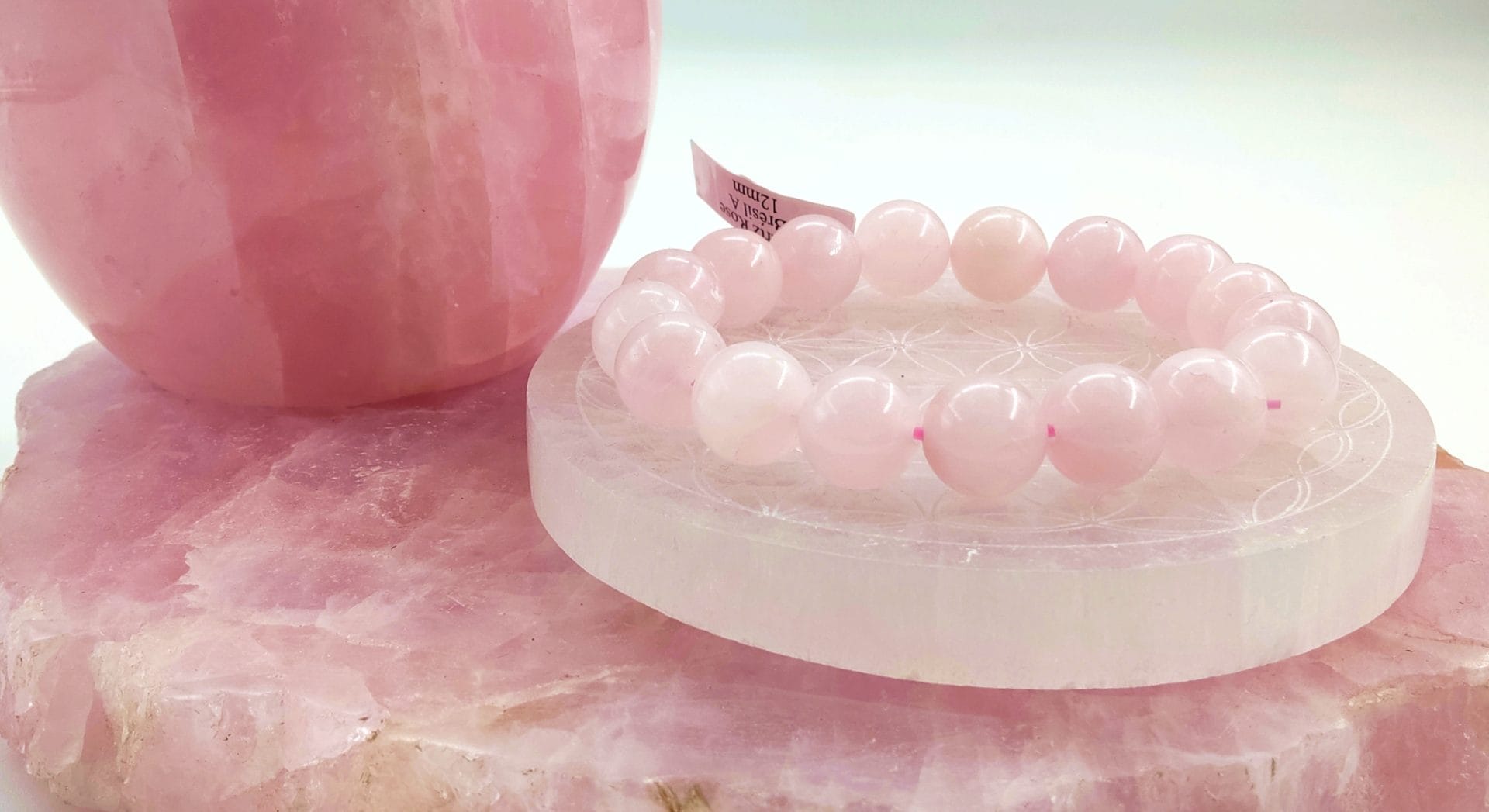 Bracelet en perles de quartz rose posé sur une plaque de sélénite gravée du motif Fleur de Vie, avec en arrière-plan une grande sphère et une base brute de quartz rose.