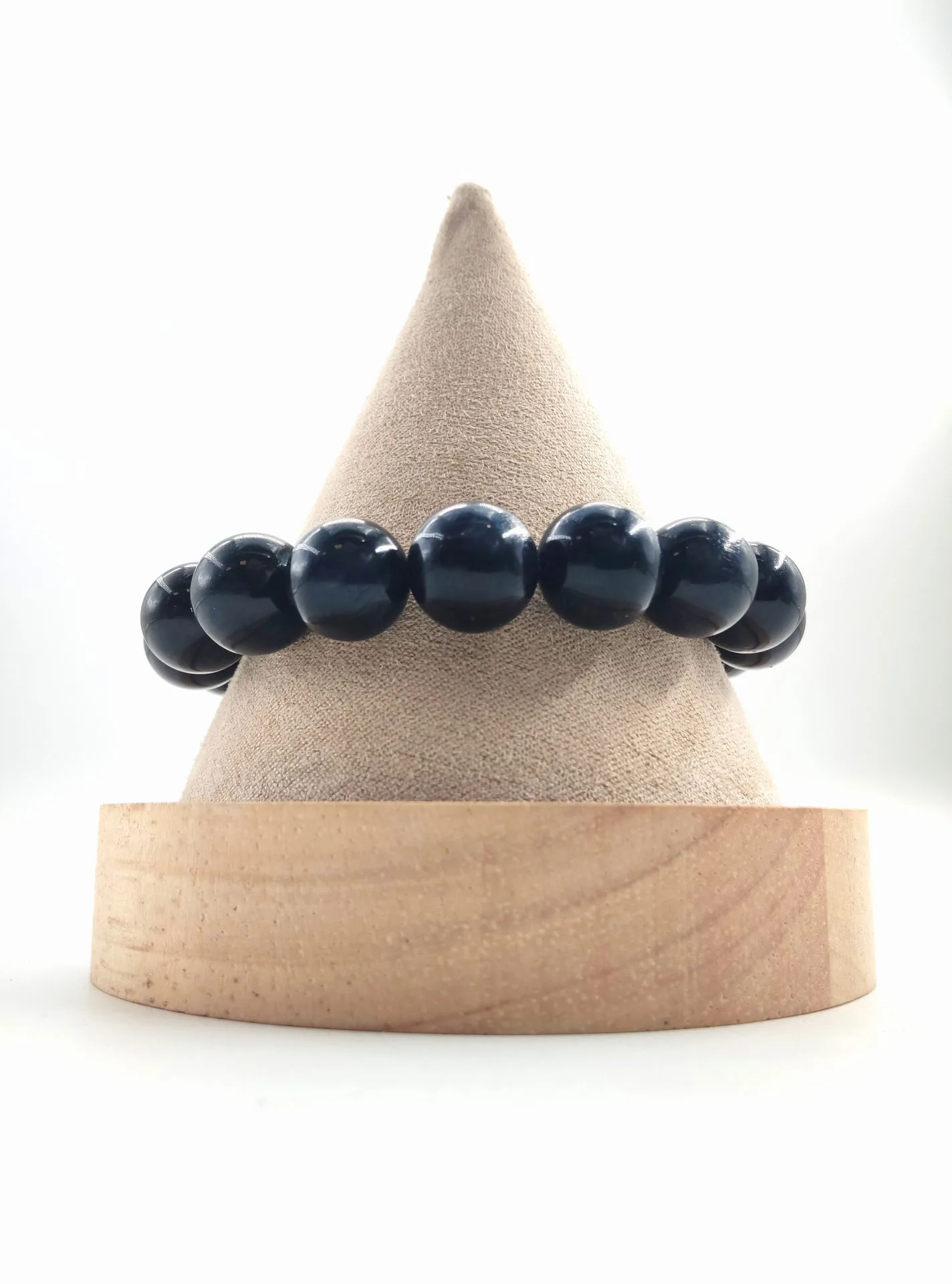 Bracelet en perles rondes d’œil de faucon, pierre sombre aux reflets bleu-noir chatoyants, présenté sur un présentoir conique en tissu beige avec socle en bois clair.