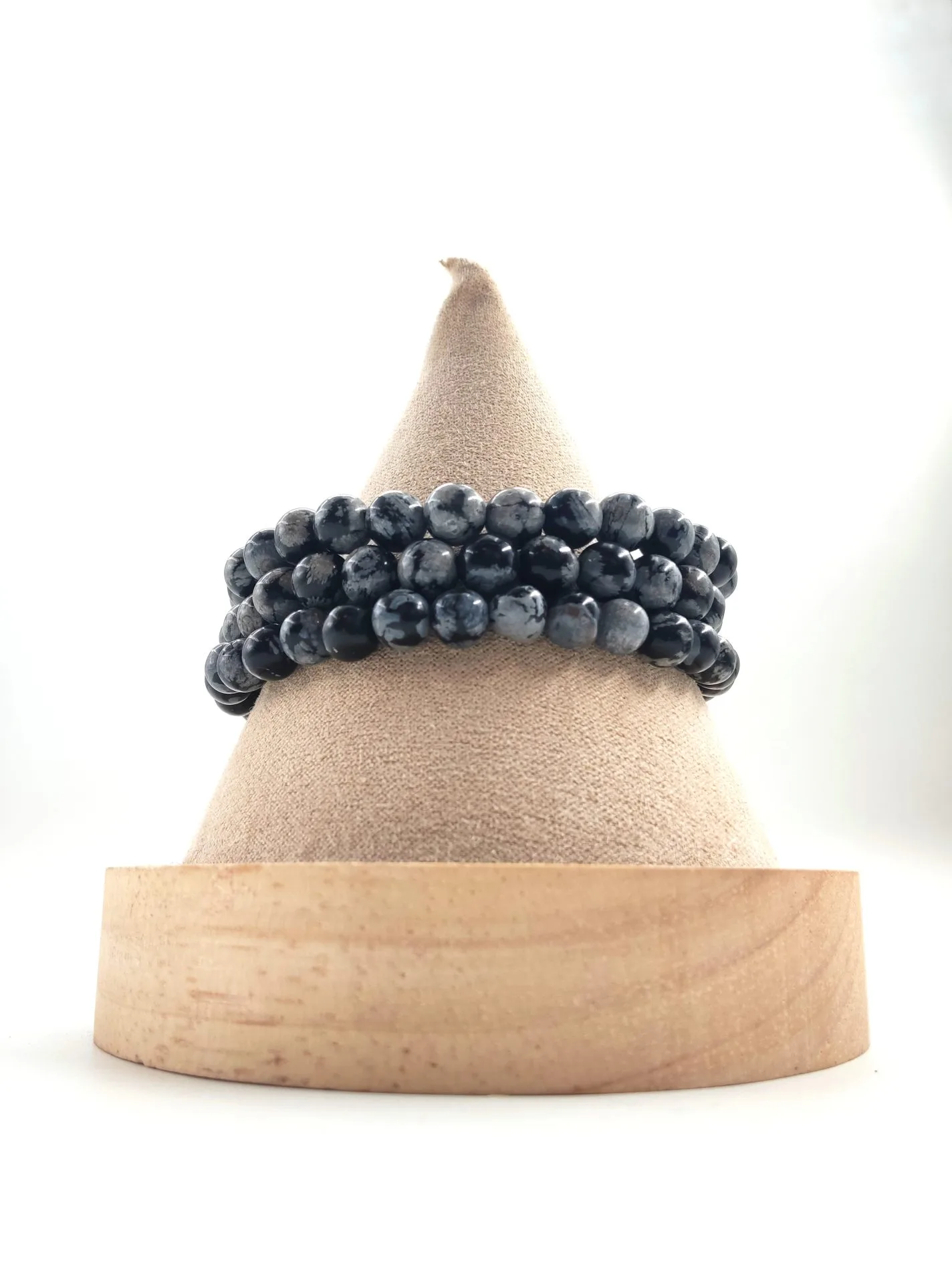 Bracelet en perles rondes d’obsidienne neige, pierre noire mouchetée de gris, présenté enroulé sur un présentoir conique en tissu beige avec socle en bois clair.