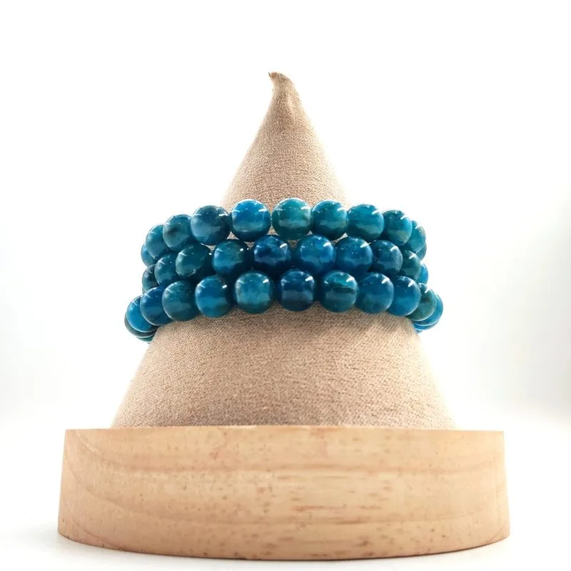 Bracelet en perles rondes de pierre naturelle bleu profond avec des reflets turquoise, présenté enroulé sur un présentoir conique en tissu beige avec socle en bois clair.