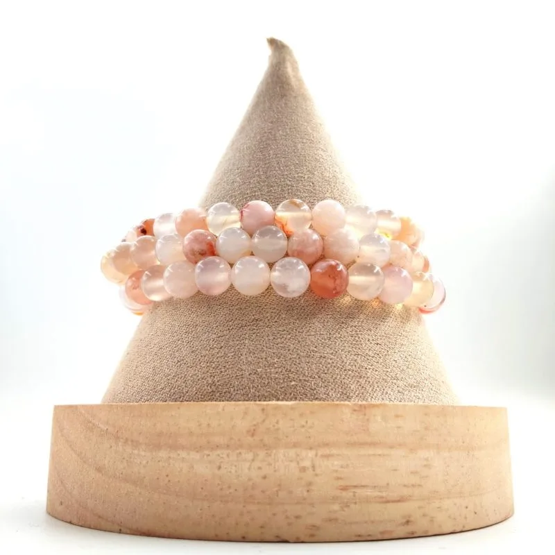 Bracelet en perles rondes de pierre naturelle aux teintes douces rose, pêche et translucides, présenté enroulé sur un présentoir conique en tissu beige avec socle en bois clair.