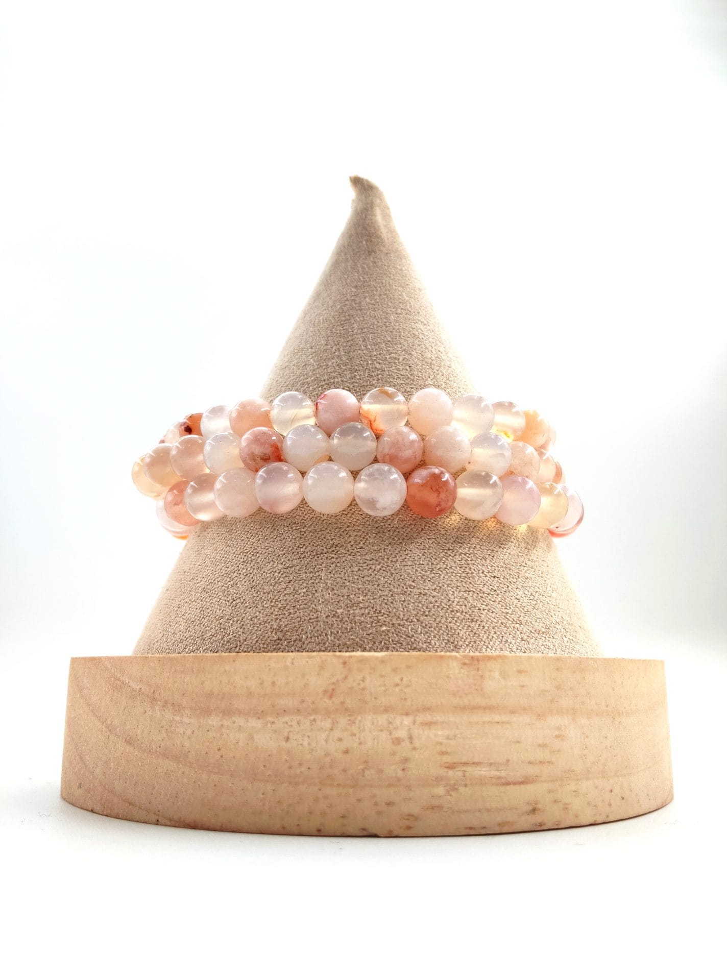 Bracelet en perles rondes de pierre naturelle aux teintes douces rose, pêche et translucides, présenté enroulé sur un présentoir conique en tissu beige avec socle en bois clair.