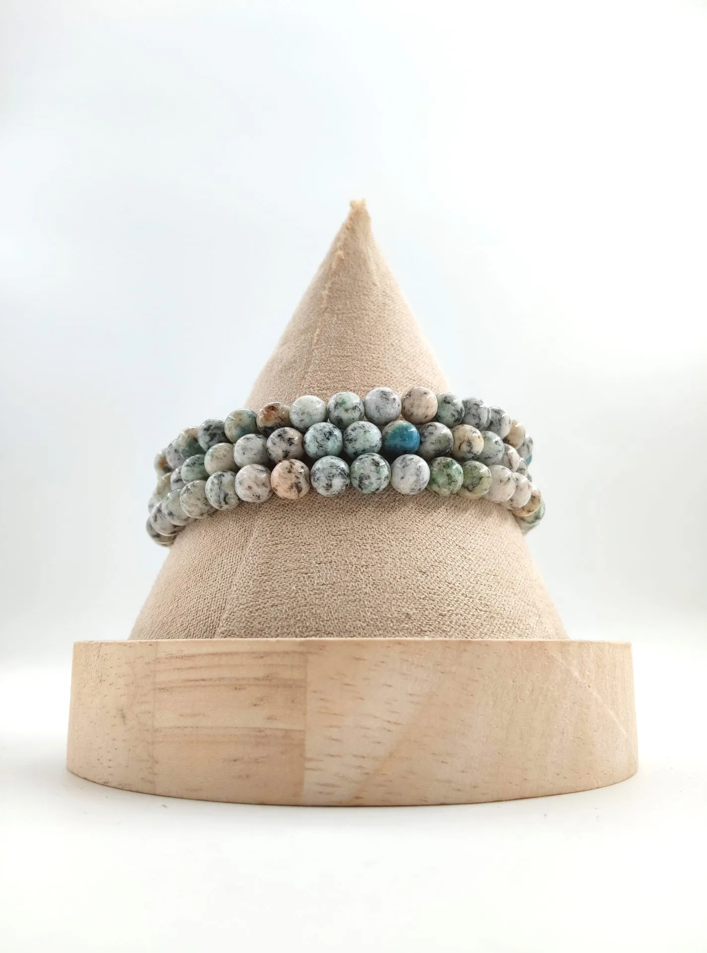 Bracelets en perles de jaspe K2, pierre blanche mouchetée de noir avec des inclusions bleues d’azurite, posés sur un présentoir conique en tissu beige avec socle en bois clair.