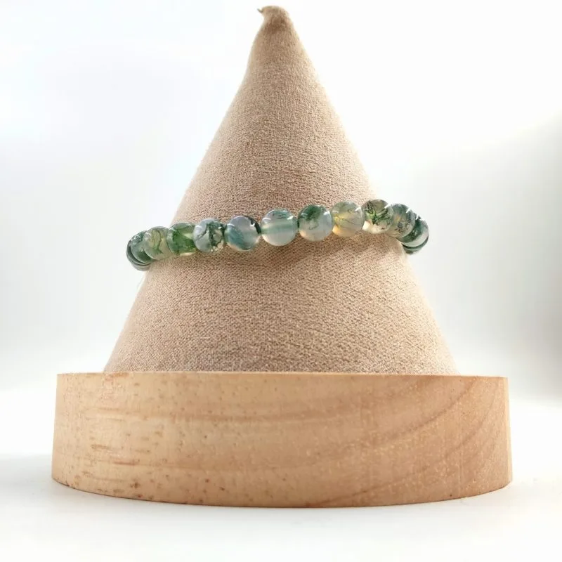 Bracelet en perles rondes d’agate mousse, aux nuances translucides blanches et vertes rappelant des inclusions végétales, présenté sur un cône en tissu beige avec base en bois clair.