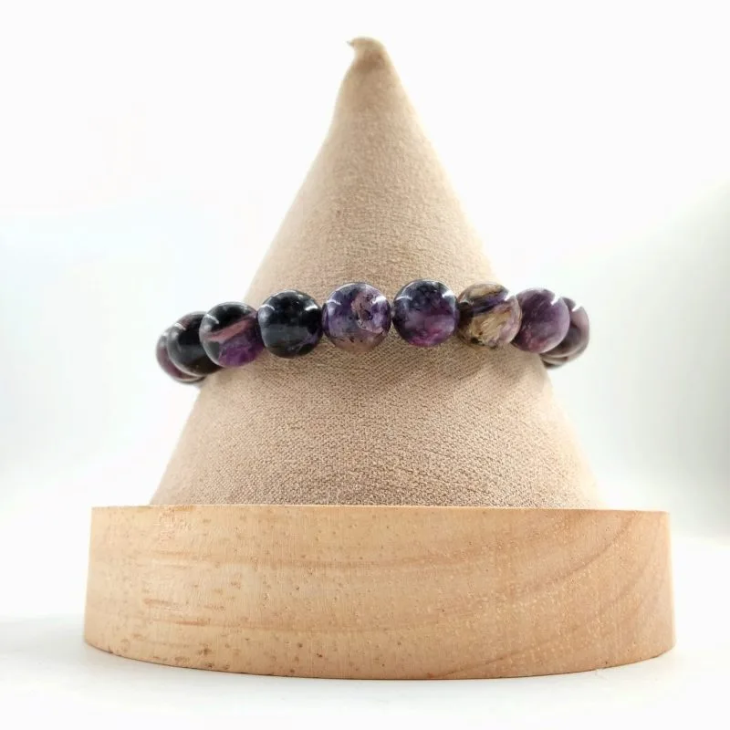 Bracelet en perles rondes de charoïte violette aux nuances noires, blanches et dorées, présenté sur un cône en tissu beige avec base en bois clair.
