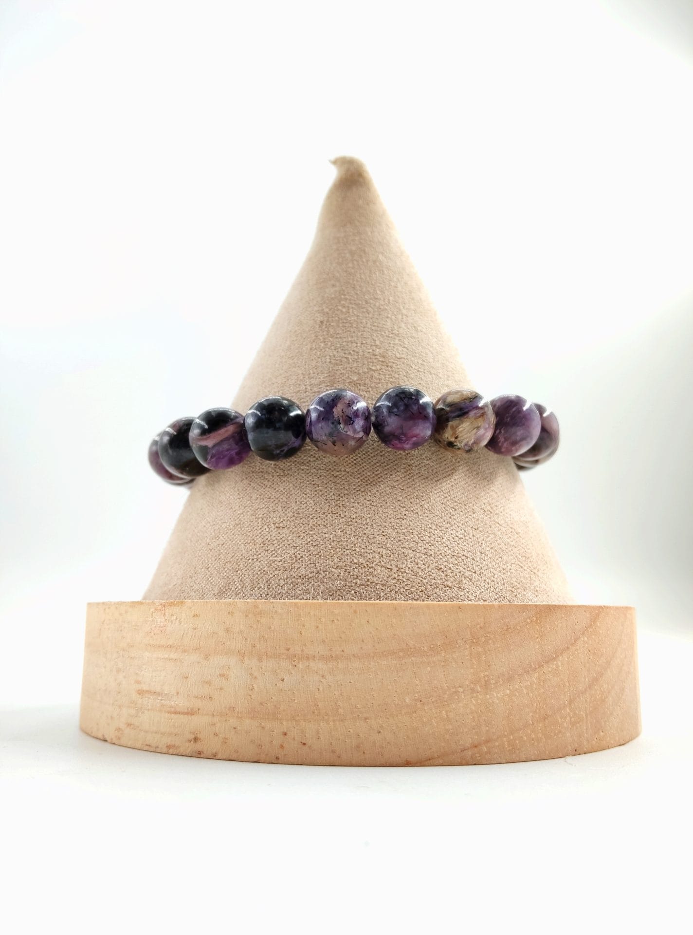 Bracelet en perles rondes de charoïte violette aux nuances noires, blanches et dorées, présenté sur un cône en tissu beige avec base en bois clair.