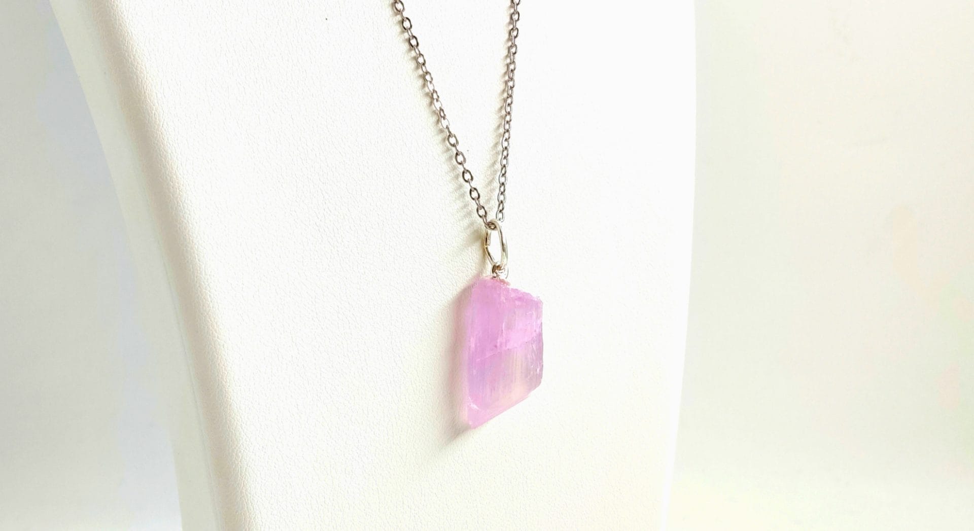 Pendentif en pierre brute de kunzite rose, monté sur une bélière argentée et suspendu à une chaîne fine en métal argenté, présenté sur un présentoir blanc.
