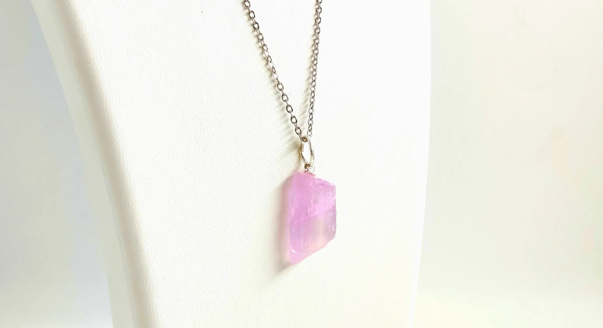 Pendentif en pierre brute de kunzite rose, monté sur une bélière argentée et suspendu à une chaîne fine en métal argenté, présenté sur un présentoir blanc.