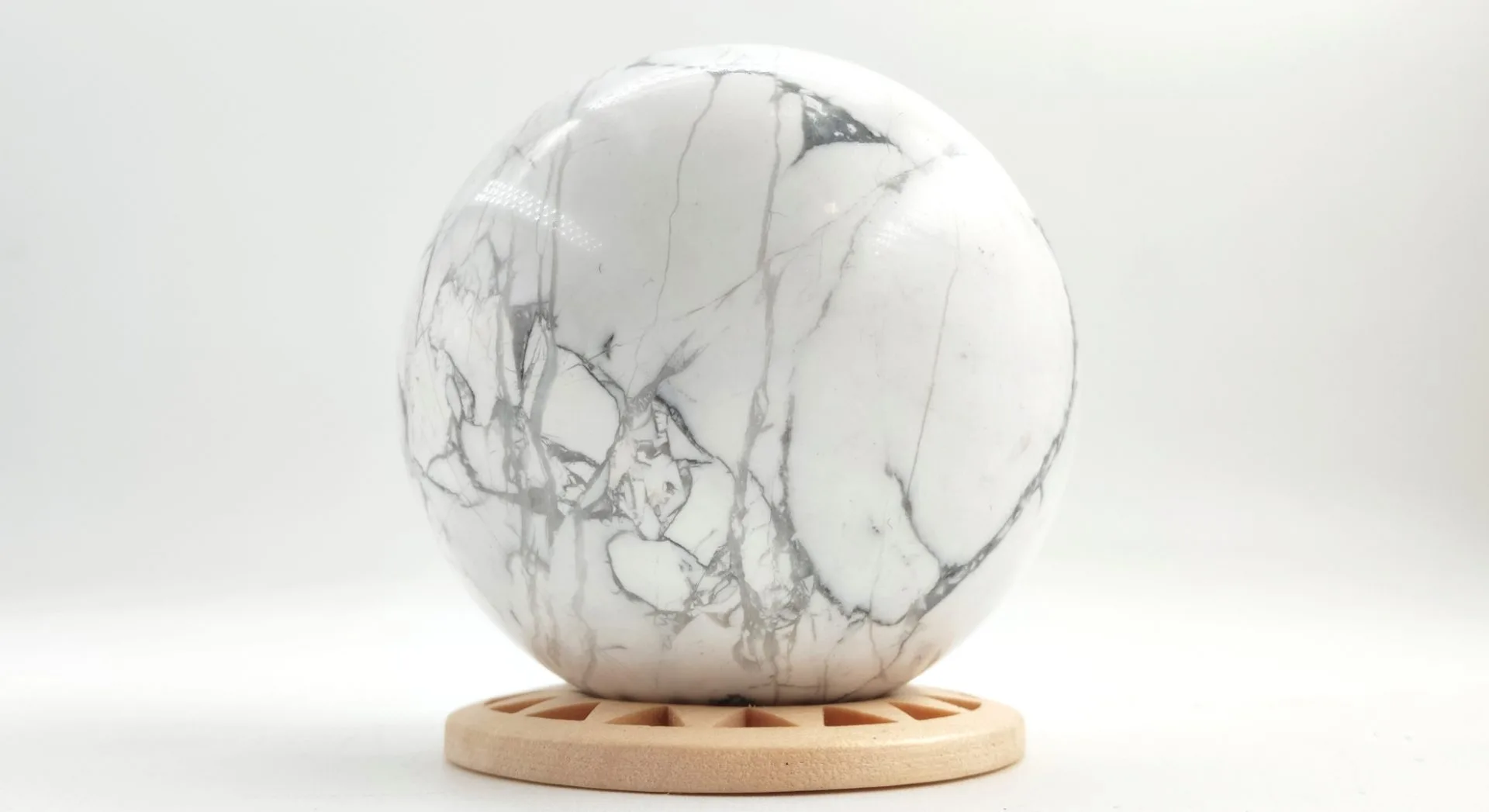Sphère en howlite blanche veinée de gris, polie et posée sur un support rond en bois clair, présentée sur fond blanc.