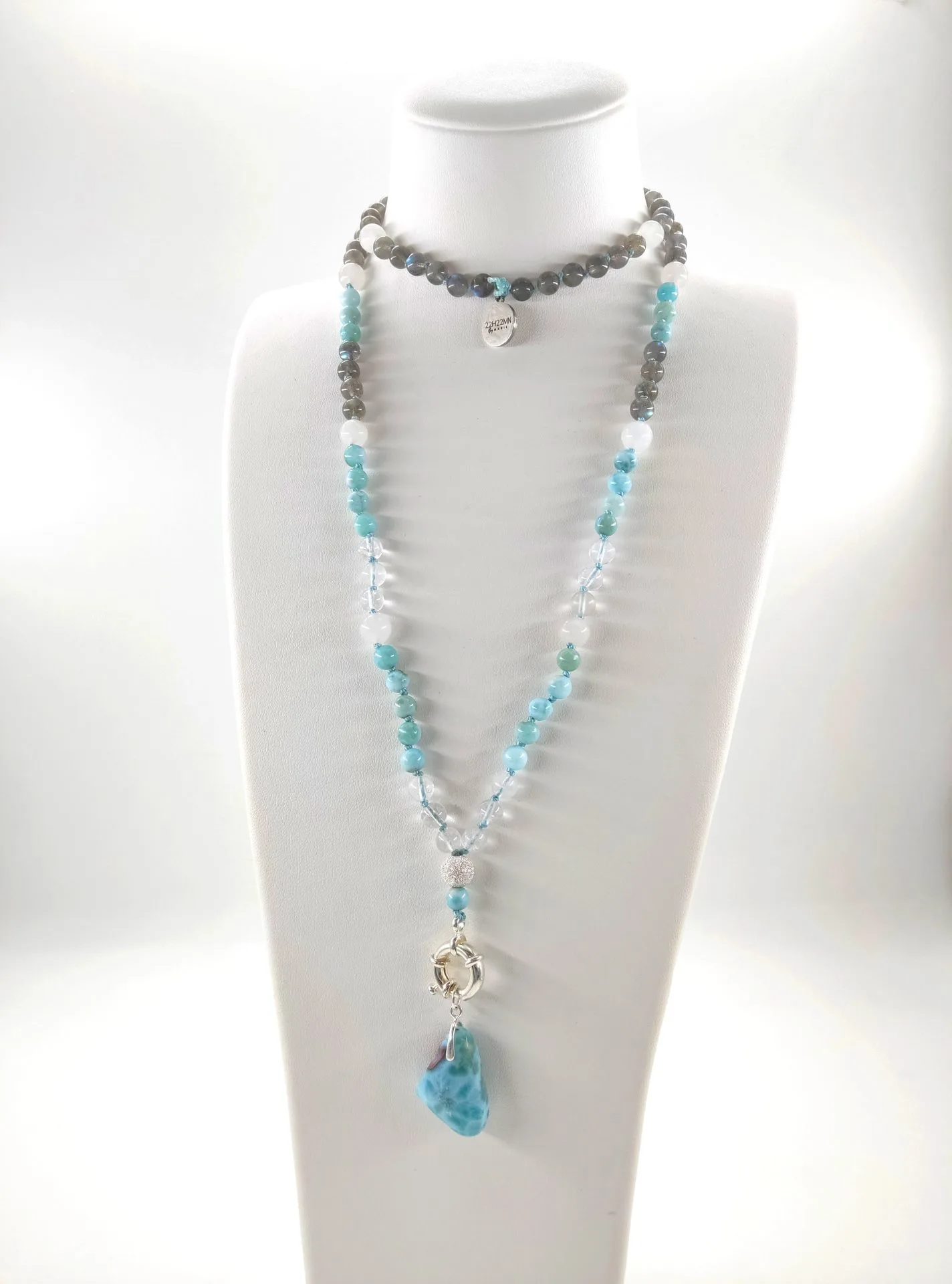 Collier long en perles bleues, blanches et grises exposé sur un présentoir blanc. Le bijou alterne de petites perles rondes brillantes dans des tons bleu turquoise, blanc laiteux et gris foncé, avec quelques perles transparentes. Au centre pend une pierre brute bleue marbrée, suspendue à un anneau argenté texturé. Le style global est lumineux, artisanal et inspiré du bien-être.