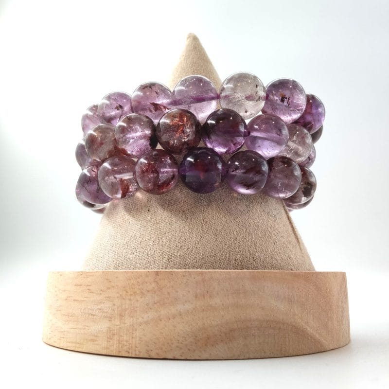 L'image présente trois bracelets de perles d'améthyste. Les perles alternent entre un violet translucide et des zones plus claires, presque cristallines. On y distingue nettement des inclusions sombres et des voiles internes qui donnent de la profondeur à la pierre. Cette variété d'améthyste, riche en détails naturels, offre un aspect plus organique et complexe que l'améthyste parfaitement uniforme.