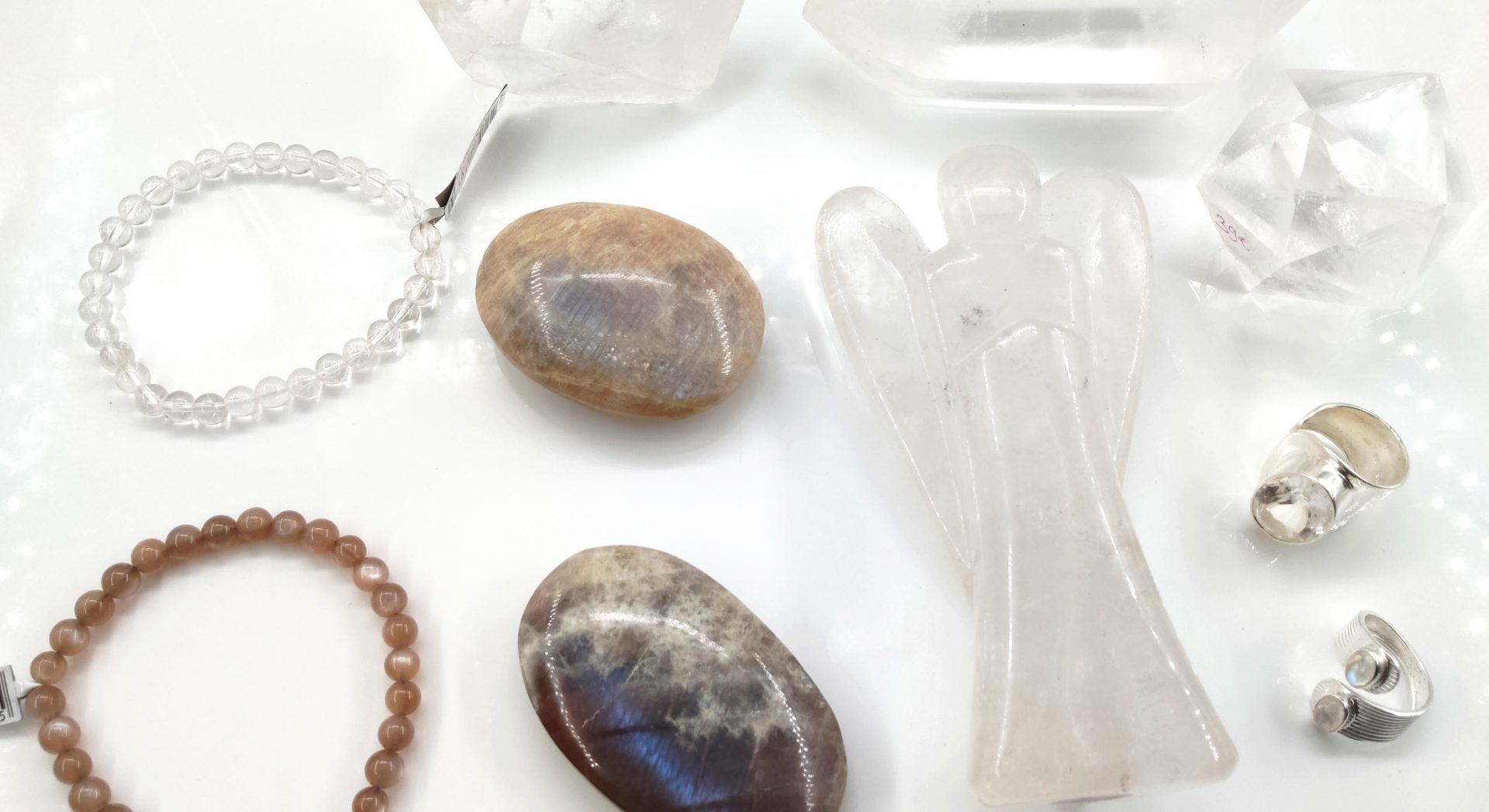 Une collection de cristaux, de bijoux et d'objets sculptés disposés sur une surface blanche unie et lumineuse. L'ensemble dégage une esthétique épurée et minérale.