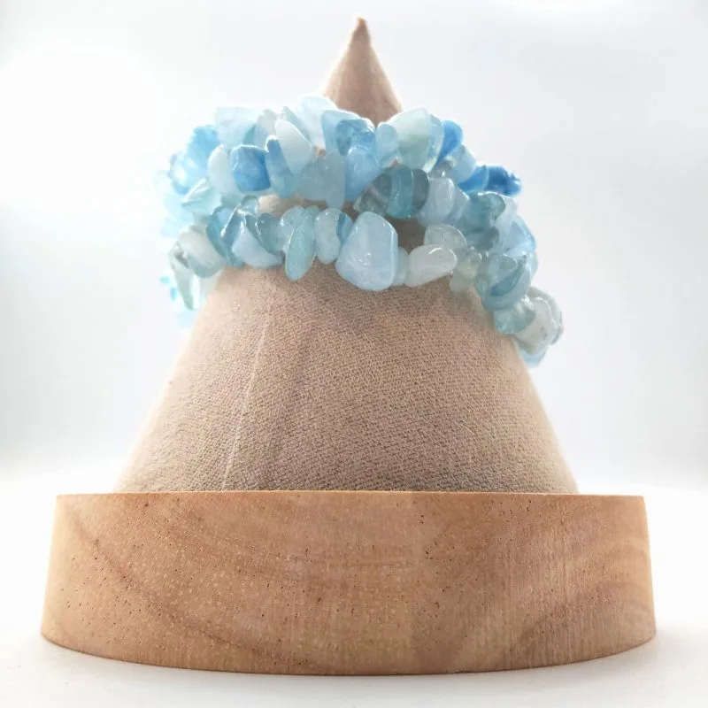 Cette photographie de studio met en lumière un bracelet artisanal en pierres naturelles d'Aigue-marine, présenté sur un support conique élégant. Le bijou se distingue par son style "baroque", composé de multiples éclats de pierres polies aux formes irrégulières et organiques, créant un relief texturé et dynamique. La palette de couleurs offre un dégradé apaisant de nuances aquatiques, allant du bleu ciel cristallin au blanc laiteux, avec quelques reflets translucides qui captent la lumière de manière diffuse.