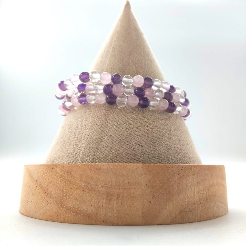 Bracelet élastique triangle d'or en lithothérapie : perles facettées alternées améthyste violette, quartz rose rose pâle et cristal de roche transparent clair (environ 6-8 mm). Posé sur un cône beige en bois clair, vue de face sur fond blanc lumineux. Harmonie émotionnelle et spirituelle pour chakras cœur et coronal.