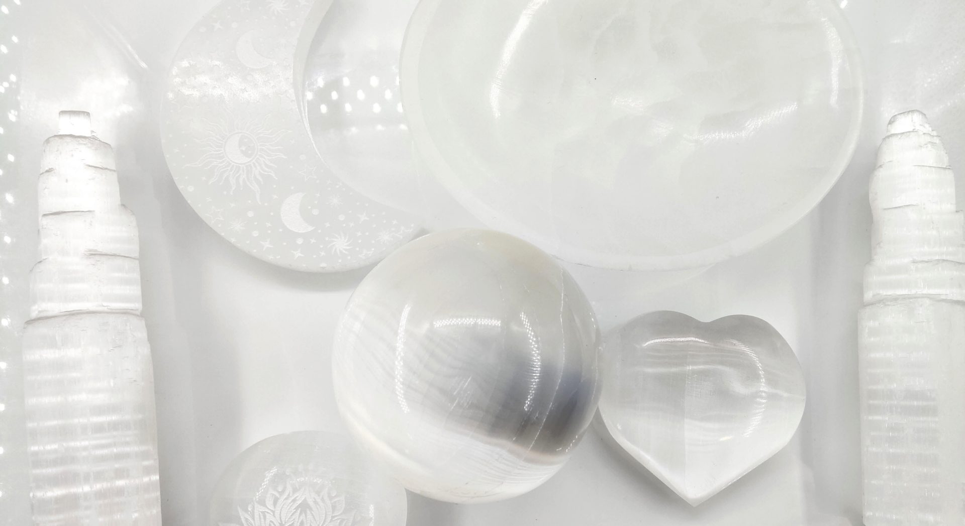 Une composition harmonieuse de plusieurs objets sculptés dans une pierre blanche satinée et translucide (sélénite), disposés sur un fond blanc lumineux. L'ensemble présente un éclat soyeux avec des reflets chatoyants linéaires.