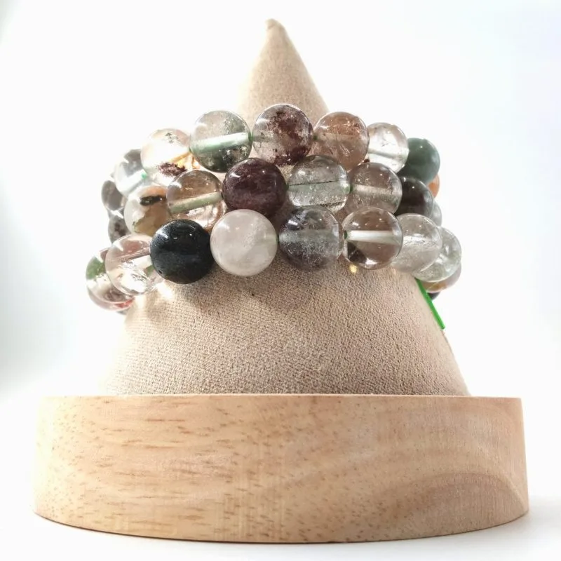 Cette image expose trois bracelets de perles de quartz transparentes contenant des inclusions minérales diverses, souvent appelées quartz fantômes. On y observe des formations internes de couleurs variées, allant du vert mousse au brun terreux et au noir, emprisonnées dans la clarté du cristal. L'alternance entre la transparence pure du quartz et ces paysages miniatures à l'intérieur de chaque sphère crée un effet visuel organique et fascinant, mis en valeur par les reflets brillants de l'éclairage frontal.