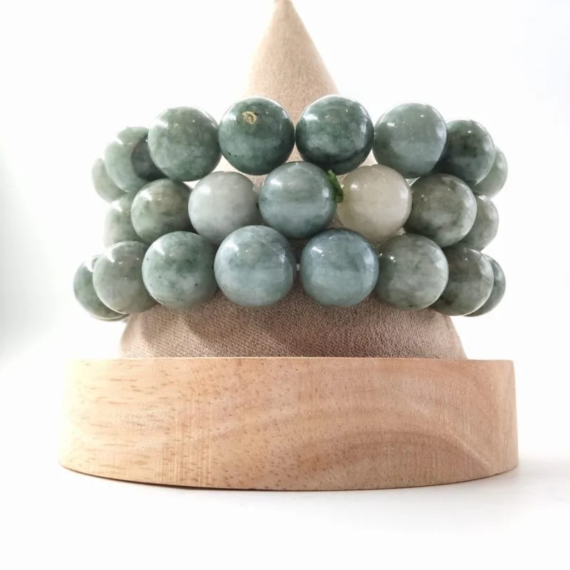 Cette image présente trois bracelets de perles de jade, disposés sur un présentoir conique en velours beige avec sa base en bois clair. Les perles affichent une teinte vert sauge mouchetée, avec des variations naturelles allant du vert d'eau très clair à un vert forêt plus soutenu. La texture de la pierre est opaque et légèrement marbrée, ce qui lui donne un aspect doux et organique. On remarque également une perle blanche distincte sur le bracelet du milieu, créant un contraste net avec le reste de l'ensemble sur le fond blanc épuré.