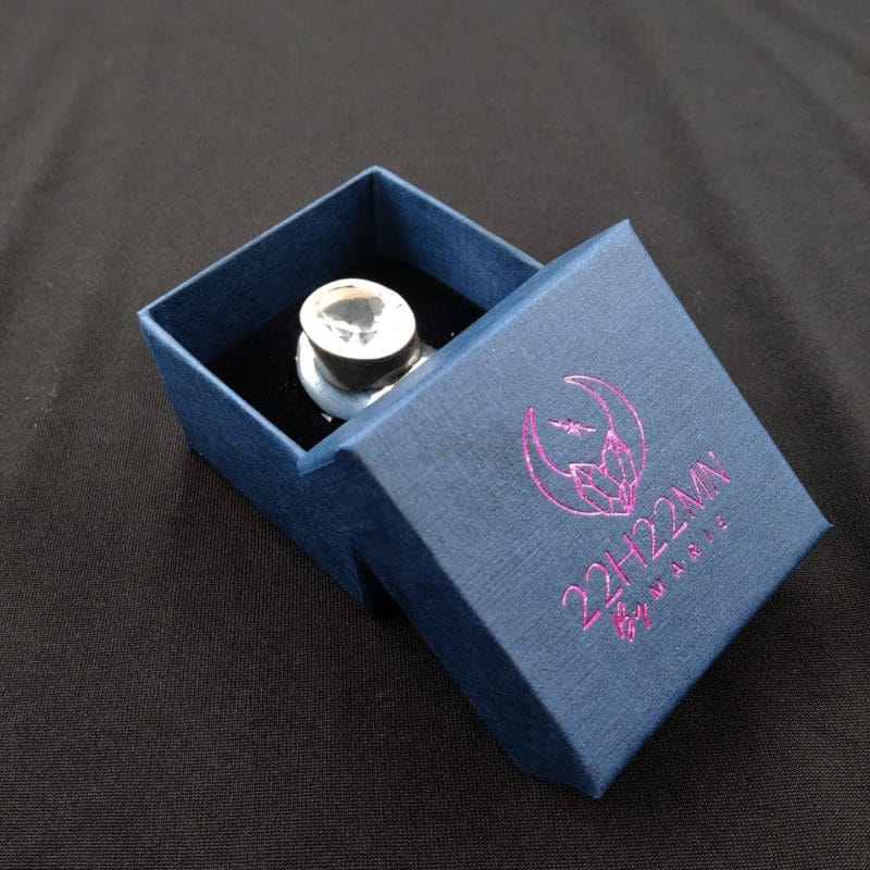 Bague ajustable en cristal de roche présentée dans son écrin bleu marine siglé 22H22MN by Marie. Le cristal de roche taillé reflète la lumière, symbolisant la clarté et l'énergie.