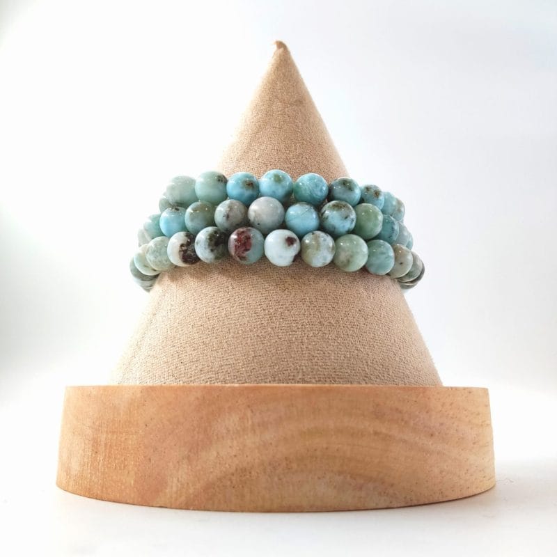 Cette image montre trois bracelets en perles superposés sur un support de présentation conique en bois et tissu. Les perles, d'un diamètre d'environ 6 mm, présentent des nuances de bleu ciel, de turquoise et de blanc crème, avec quelques inclusions brunes et grisâtres.