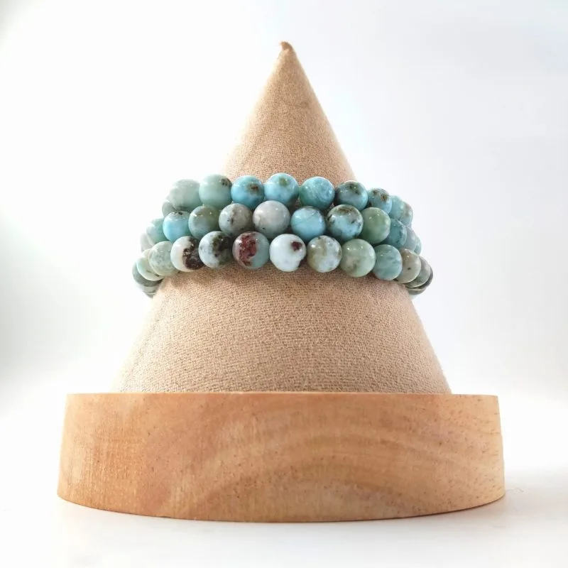 Cette image montre trois bracelets en perles superposés sur un support de présentation conique en bois et tissu. Les perles, d'un diamètre d'environ 6 mm, présentent des nuances de bleu ciel, de turquoise et de blanc crème, avec quelques inclusions brunes et grisâtres.