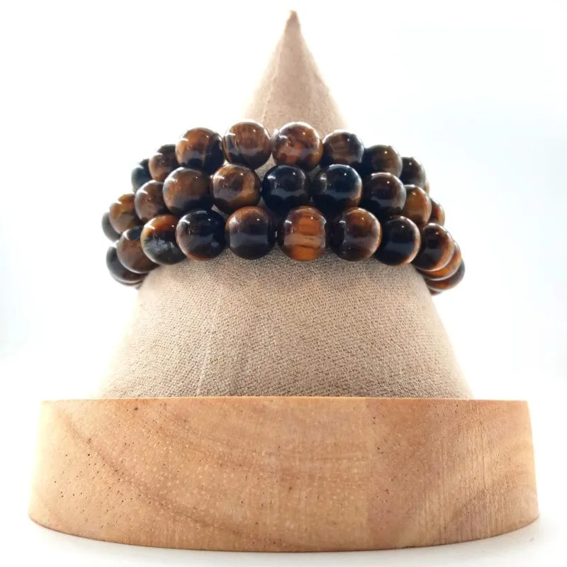 Bracelet élastique œil de tigre lithothérapie : multiples rangs de perles rondes brun doré chatoyantes (environ 8-10 mm), effet tigre rayé. Posé sur cône beige en velours, sur socle bois clair, vue de face sur fond blanc lumineux. Protection et confiance chakra solaire.